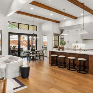 Open-concept kitchen and living area featuring Placeholder Product 1 with wooden beams, an island with stools, a dining table, and light furniture. Large windows ensure plenty of natural light.
